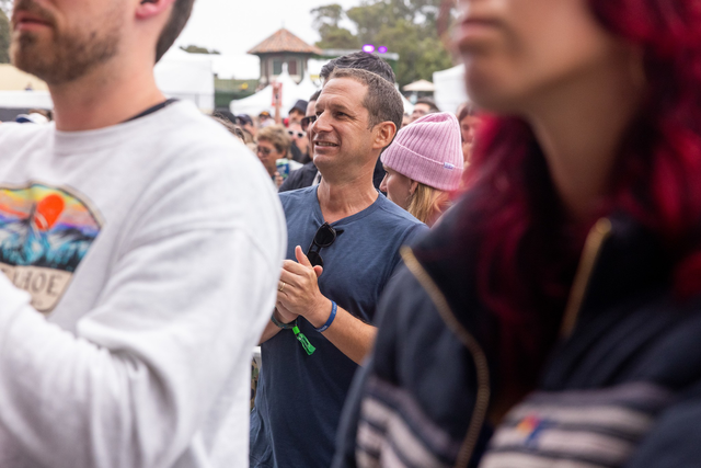 A smiling man in a blue shirt claps his hands among a crowd, with people wearing casual clothing and some with hats in an outdoor setting.